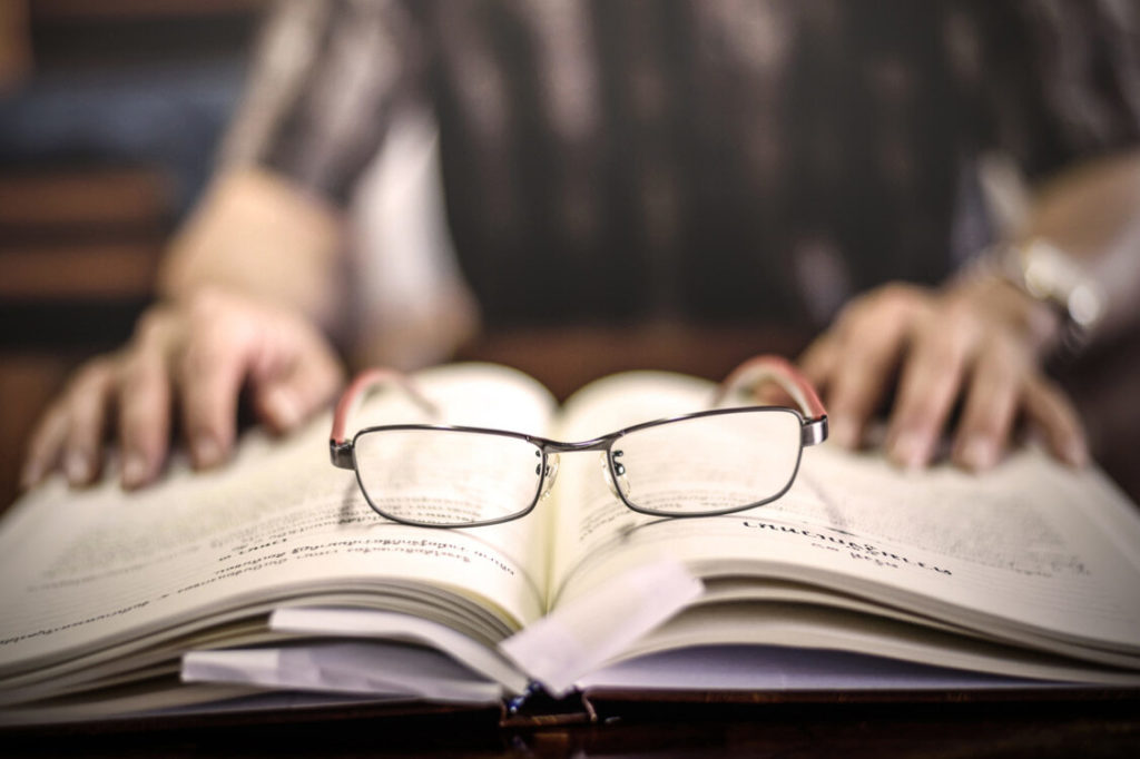 A pair of glasses resting on an open book with a person blurred out in the background