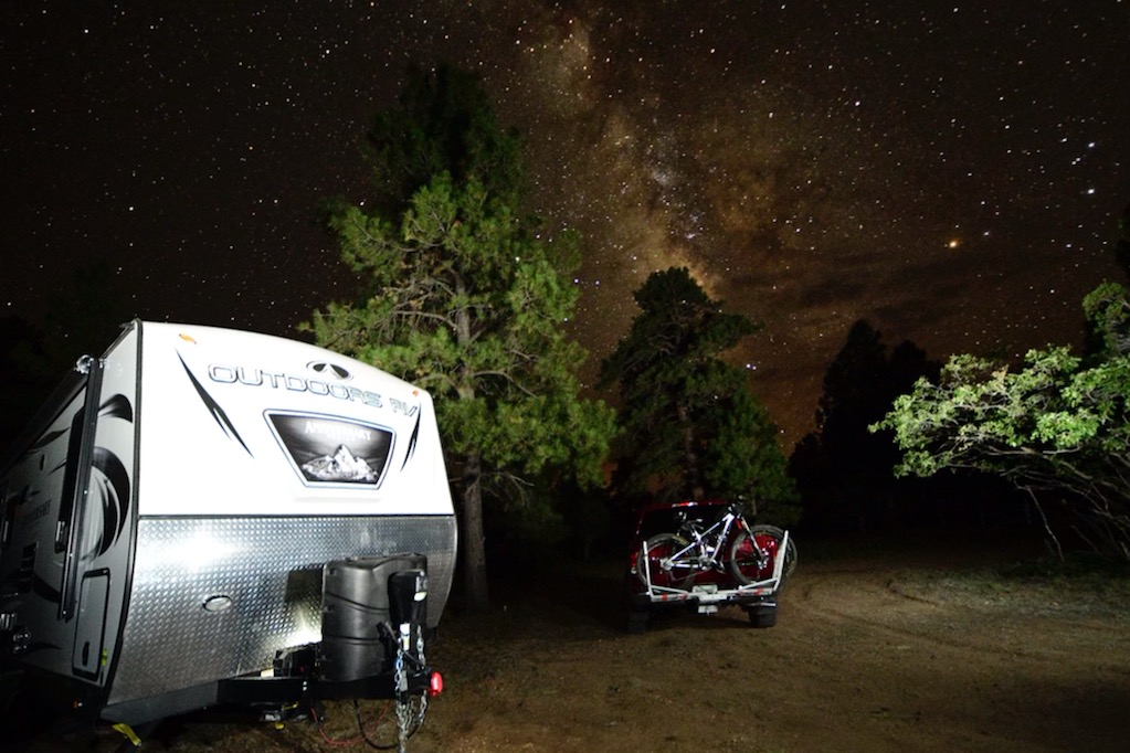 A photo of a campsite with a trailer and bikes on a bike rack. The Milky Way Galaxy is visible above the trees.