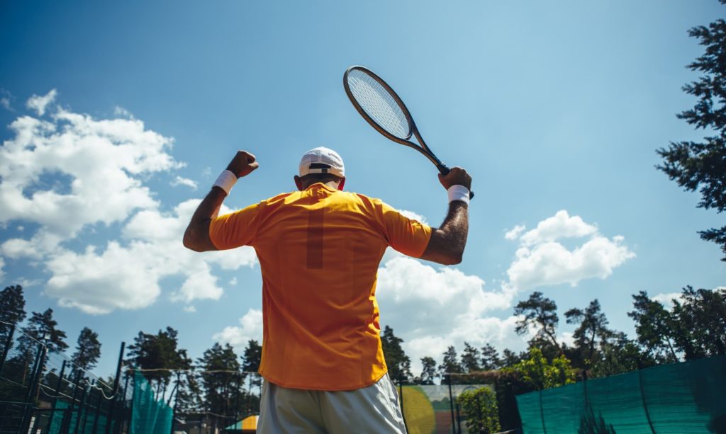 Male flourishing hands with sport equipment while turning back to camera. He winning during game while locating under blue sky