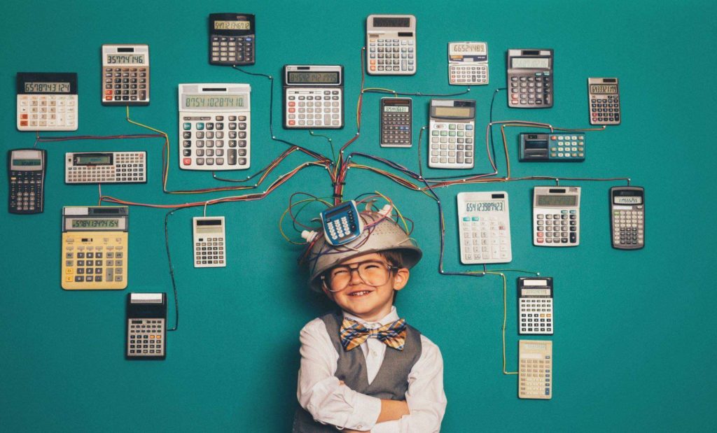 A young boy dressed in a nerdy style of clothing representing that he is numbers whiz. He is wearing a makeshift helmet that has wires coming out leading to many calculators mounted on a wall.