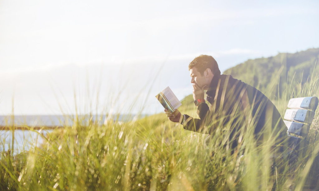 man sitting on a bench reading a book in a grassy field near the ocean