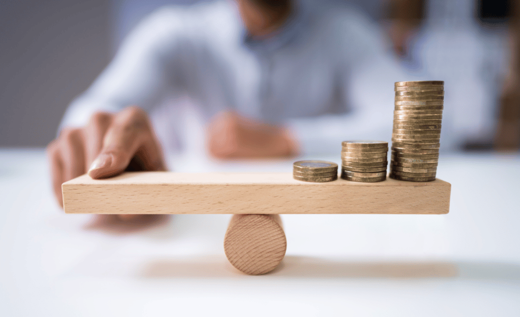 Close-up of a businessman, blurred out in the background, balancing the coin stack on wooden seesaw over desk.