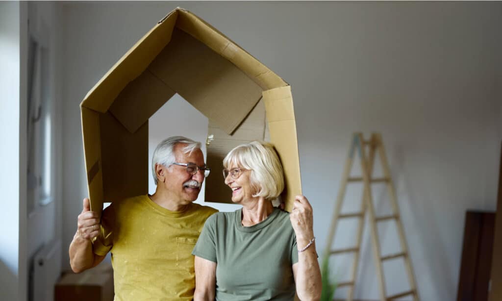 older couple holding a box over their heads shaped to look like a house while doing home renovations