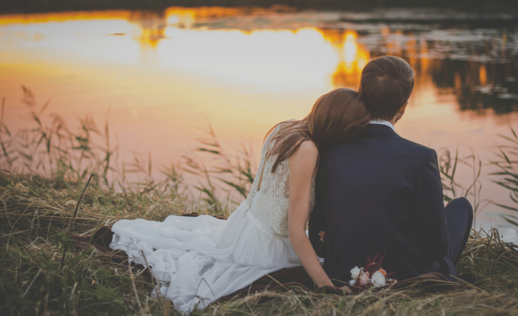 sitting woman leaning on sitting man's shoulder facing lake during golden hour