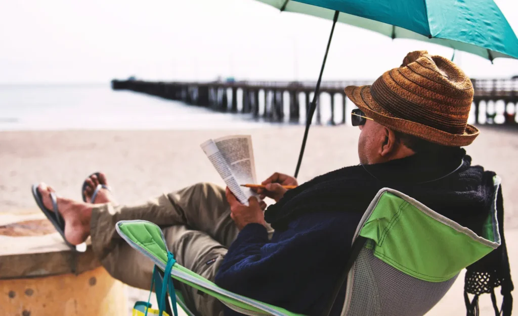 a man sitting in a chair with an umbrella by a beach reading a book and marking it pencil