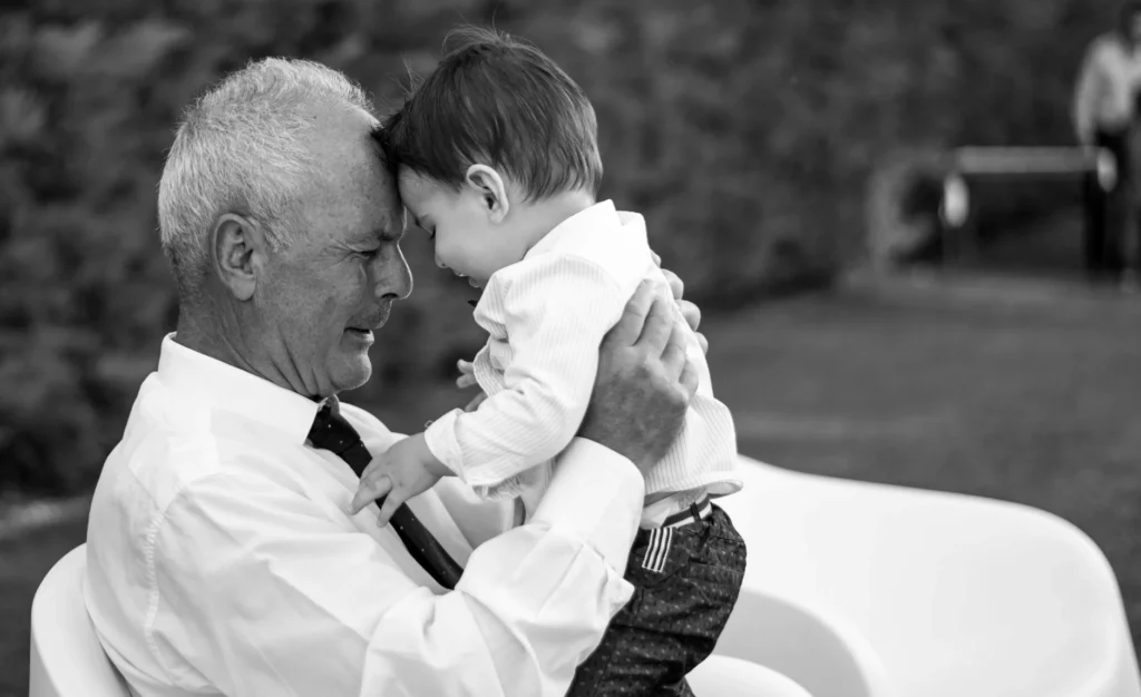 a grandfather sitting down holding his grandson on his lap. their heads are gently pressed together.