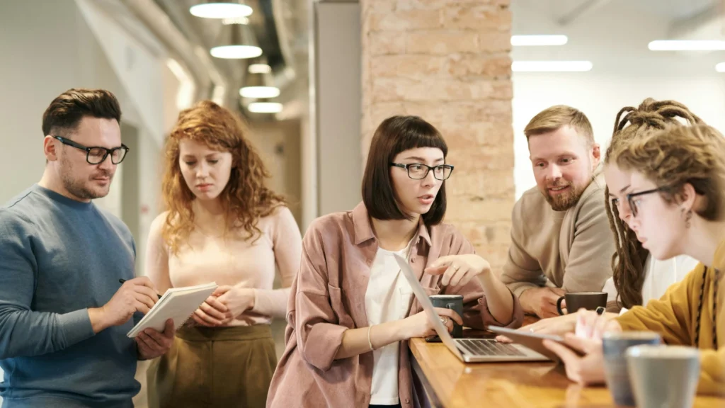 young people in an office setting working together. most are gathered around a laptop and some are gathered around a phone.